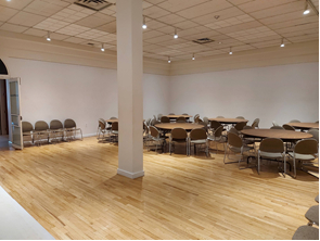 white walled room with pale wooden floors set up with round tables and grey chairs around them, and a pillar in the room